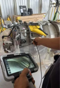 A technician uses a handheld industrial video borescope to inspect the interior of an aircraft turbine engine in a maintenance workshop, live internal footage displayed on the tablet controller