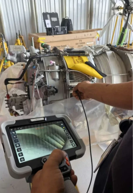 A technician uses a handheld industrial video borescope to inspect the interior of an aircraft turbine engine in a maintenance workshop, live internal footage displayed on the tablet controller