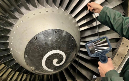 Maintenance technician holding handheld industrial borescope controller, inserting flexible probe into commercial aircraft turbofan engine front hub to inspect internal turbine blades