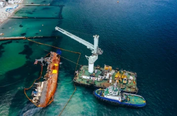 Aerial view of offshore salvage operation, crane barge and tugboat beside sunken rusted vessel with oil containment booms near coastline